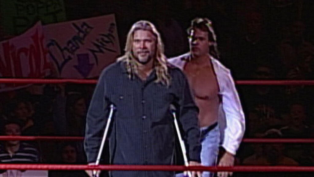 Mike Awesome - with jeans, fannypack, and white jacket - stalks Kevin Nash, who is on crutches, staring at the camera.